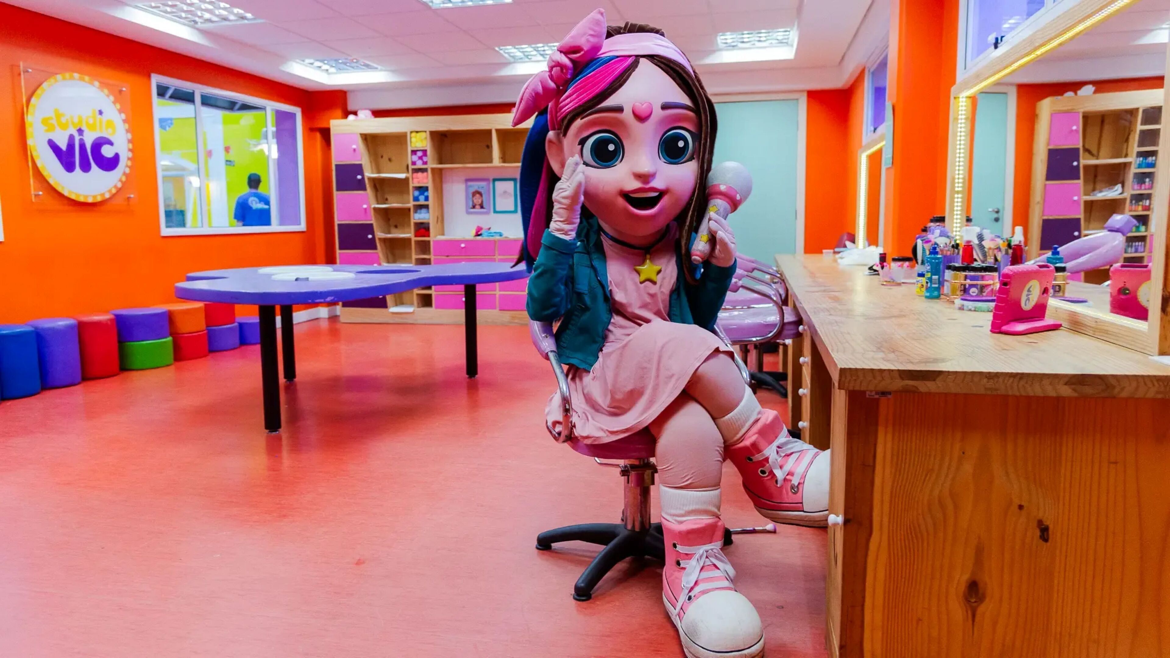 A brightly colored room with a large table, colorful storage units, and a mascot.