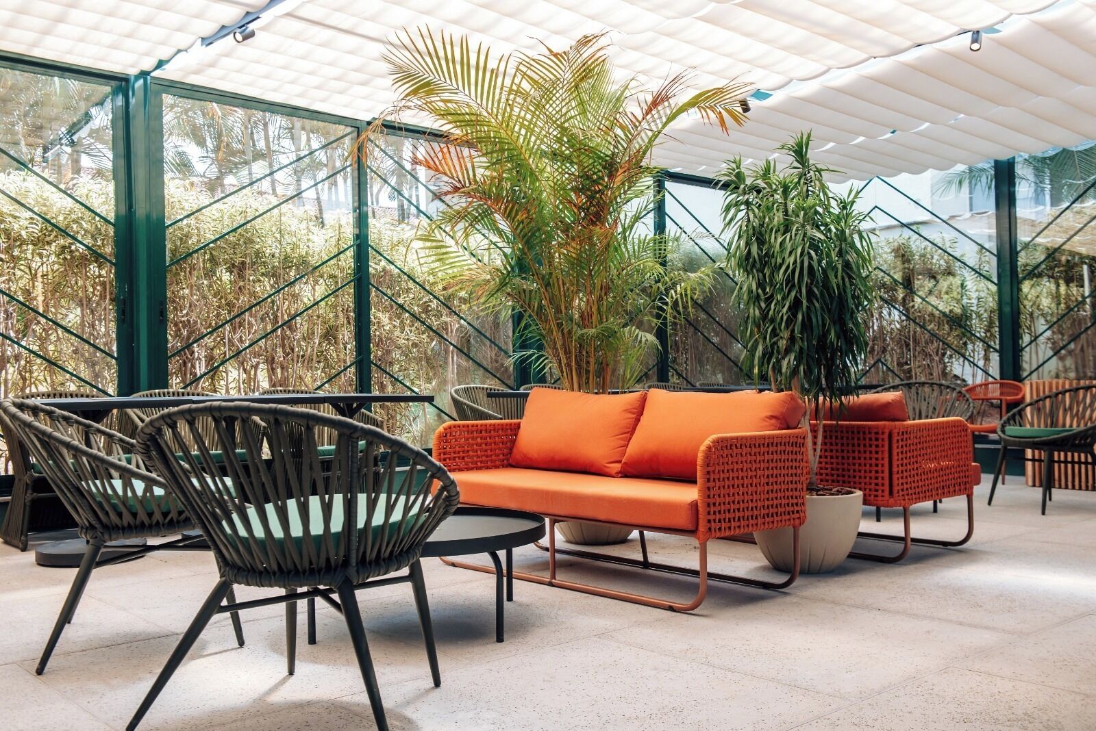 A modern outdoor seating area with wicker furniture and a bright orange sofa, surrounded by lush greenery and a glass ceiling structure.