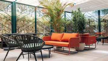 A modern outdoor seating area with wicker furniture and a bright orange sofa, surrounded by lush greenery and a glass ceiling structure.