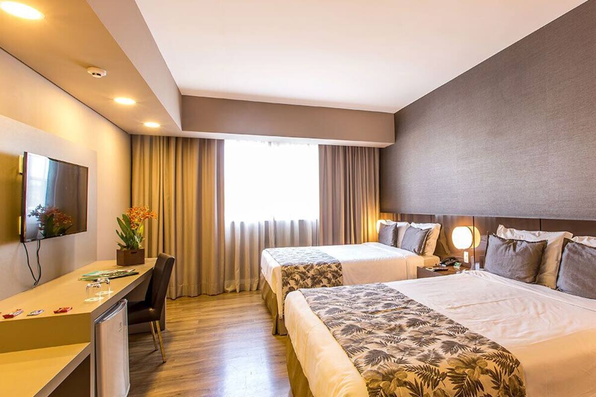 A hotel room with two beds, a desk, a flat-screen TV, and a large window with curtains.