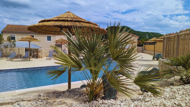 Una piscina con sillones de descanso y sombrillas, rodeada de palmeras y un edificio con techo de paja.