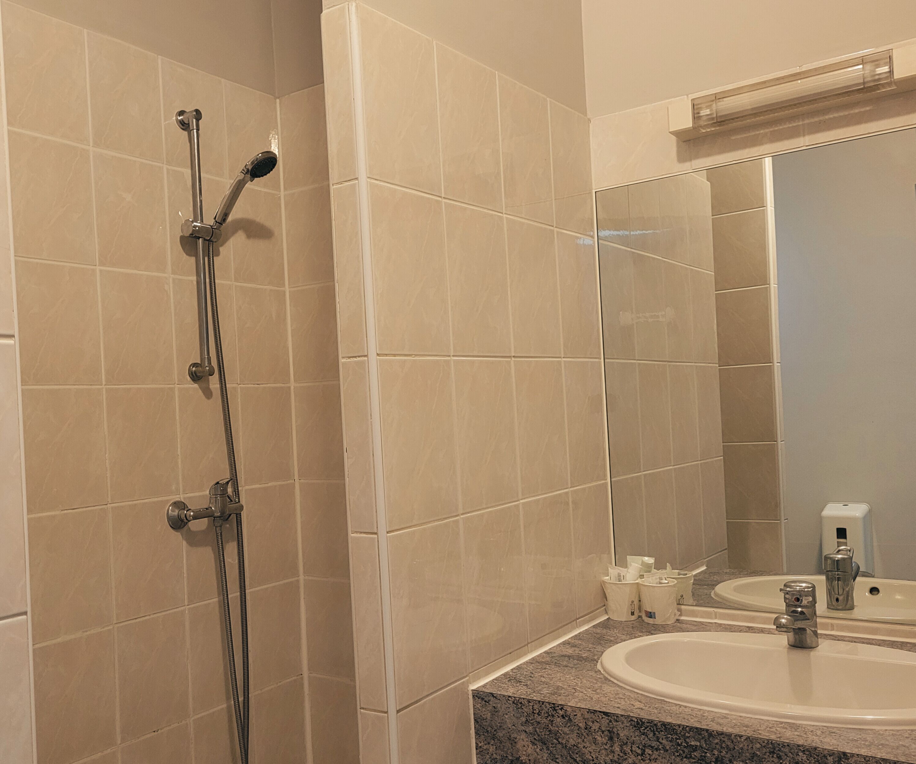 A bathroom with beige tiles, a shower with a handheld showerhead, a sink with a mirror, and a soap dispenser.