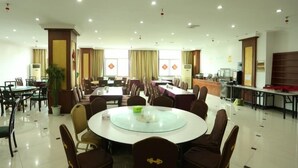 Ampla sala de jantar de hotel, com várias mesas e cadeiras, uma mesa redonda central e um balcão de buffet.
