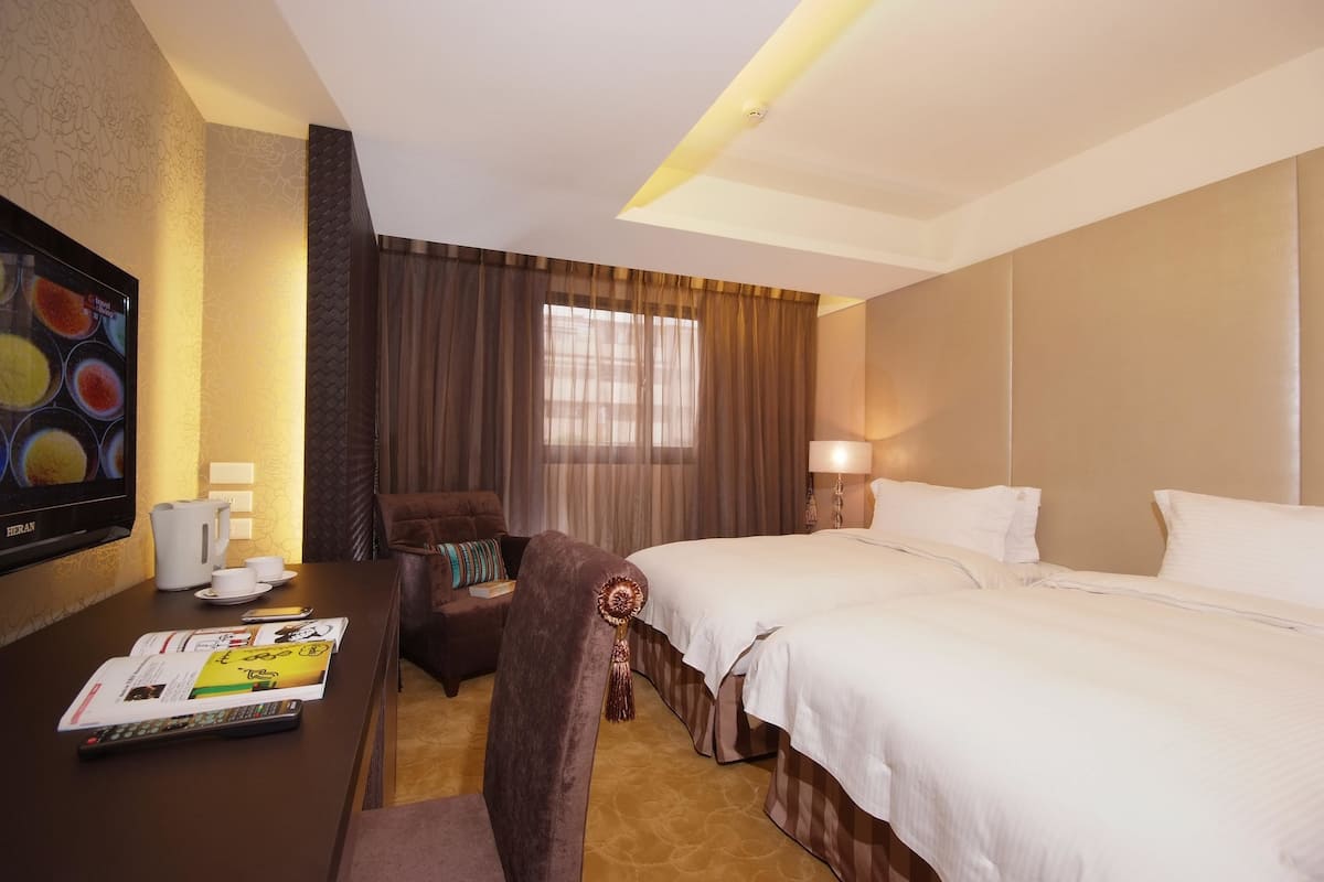A hotel room with a large bed, a desk with a remote control, a chair, a television, and a window with curtains.