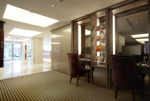 Een moderne hotellobby met een glazen scheidingswand, een receptiebalie en een zithoek met twee stoelen.