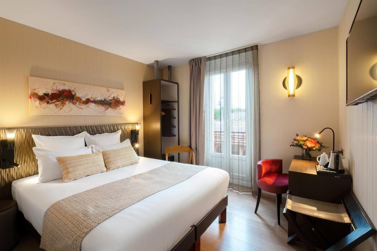 Ein Hotelzimmer mit einem großen Bett, einem Schreibtisch mit Stuhl, einem an der Wand montierten Fernseher und einem Fenster mit Vorhängen.