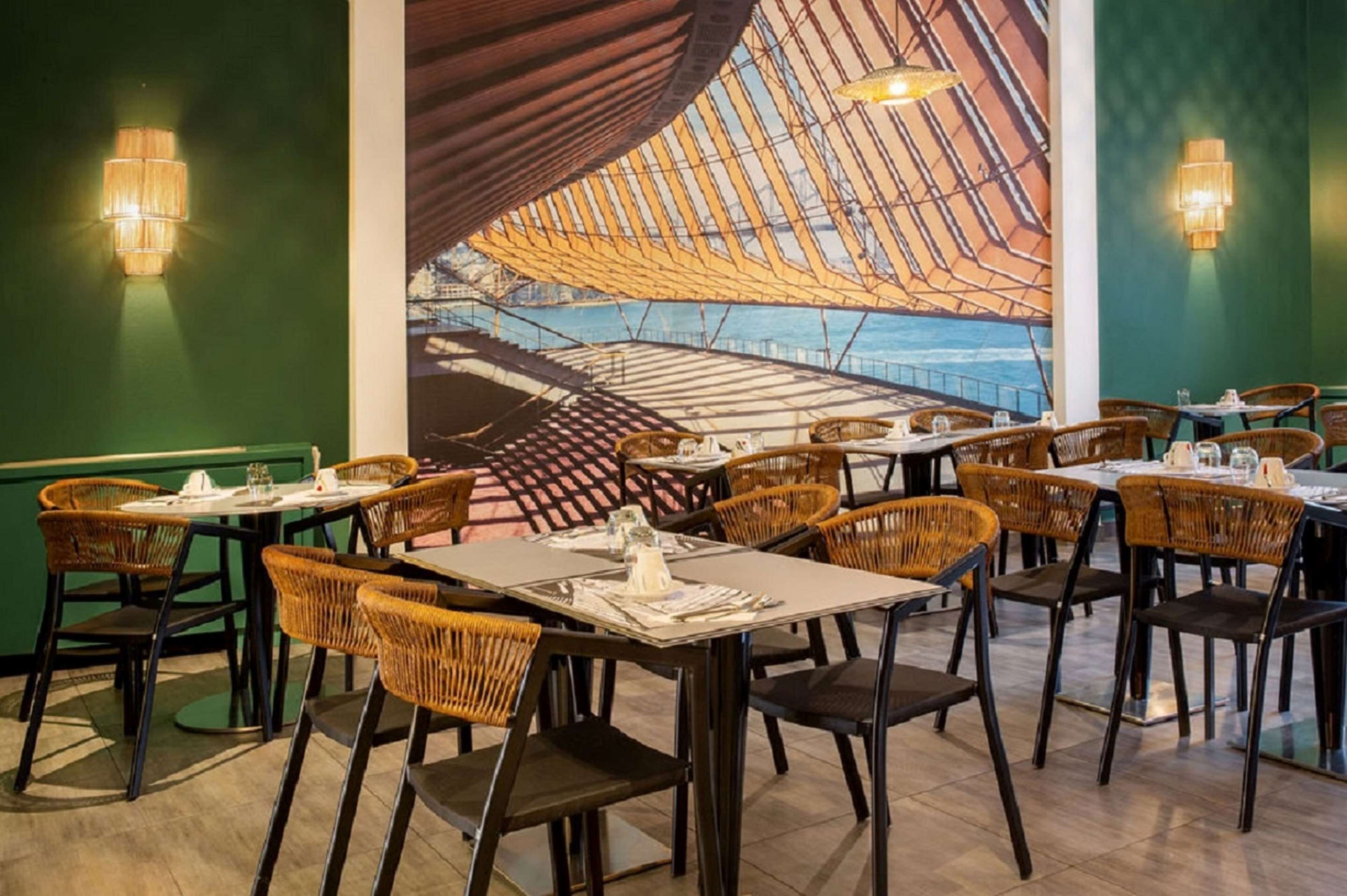 A restaurant with wicker chairs and tables, a large mural of a ship on the wall, and a green and brown color scheme.