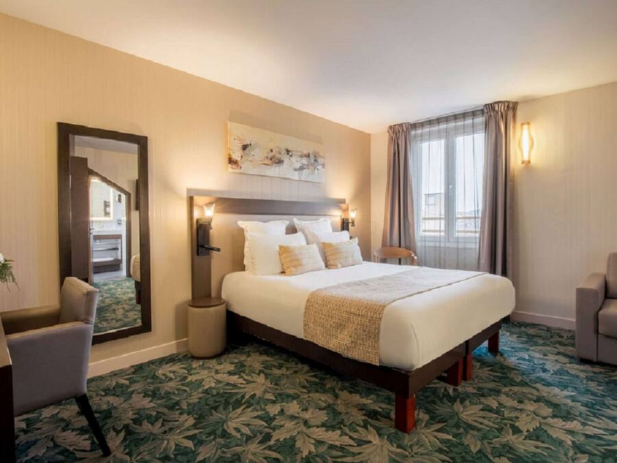 Une chambre d’hôtel avec un grand lit, une chaise, une petite table avec un vase, un miroir et une fenêtre avec des rideaux.