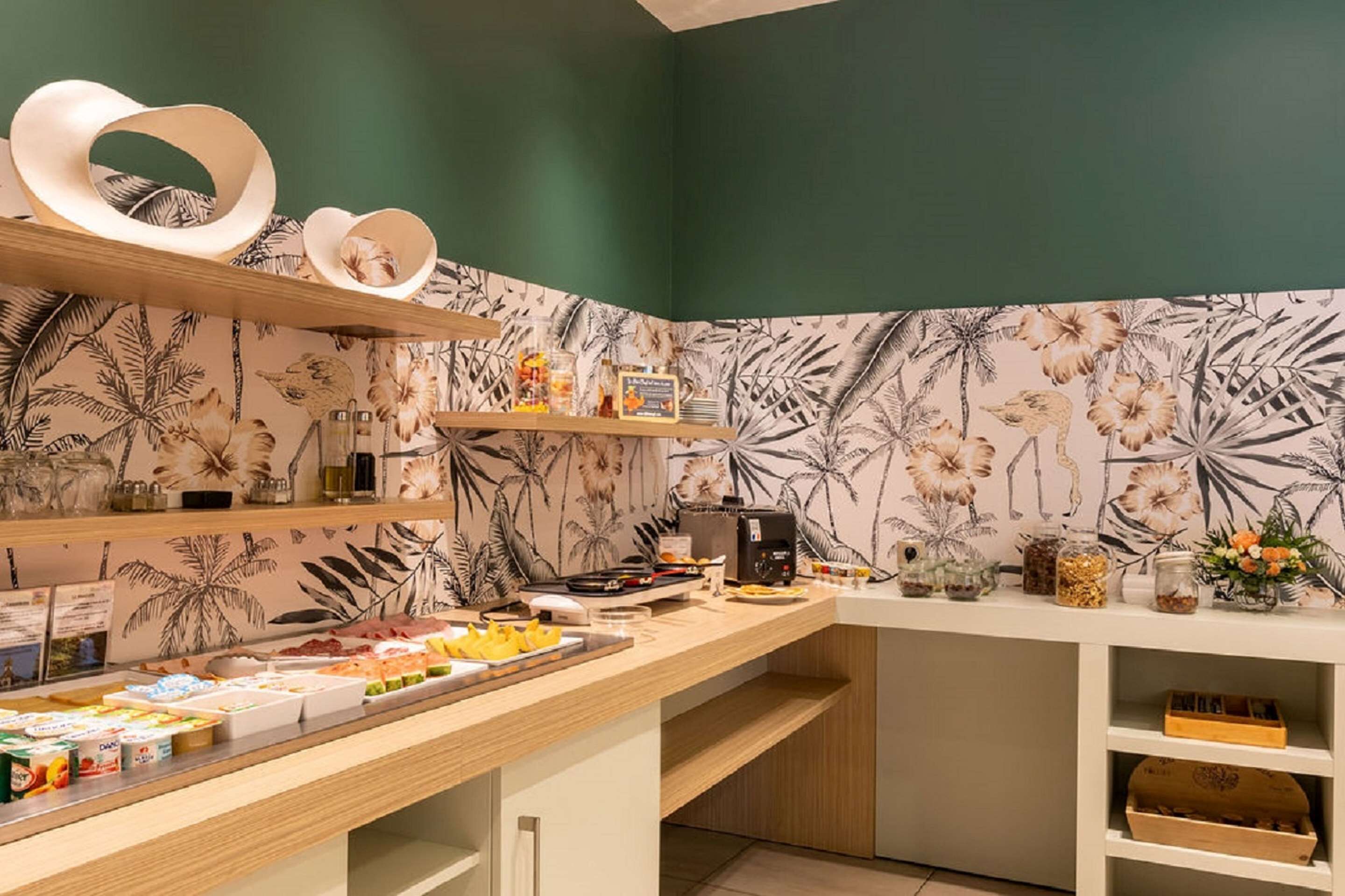 Una barra de desayuno con diferentes alimentos y condimentos, una máquina de café y elementos decorativos en estantes frente a un fondo de papel tapiz tropical.