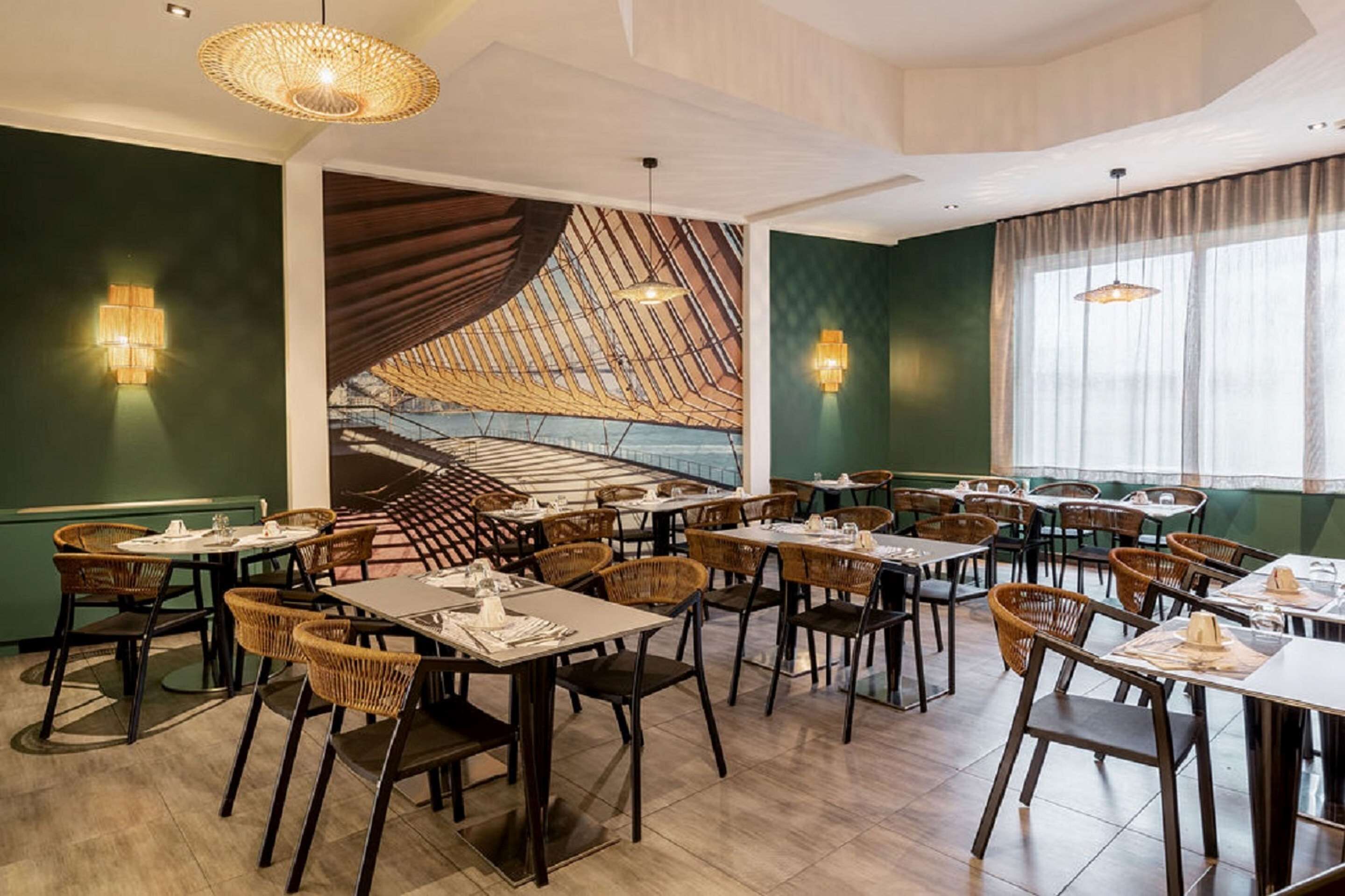 Un restaurante con una decoración moderna, que presenta un mural grande, mobiliario de mimbre e iluminación colgante.