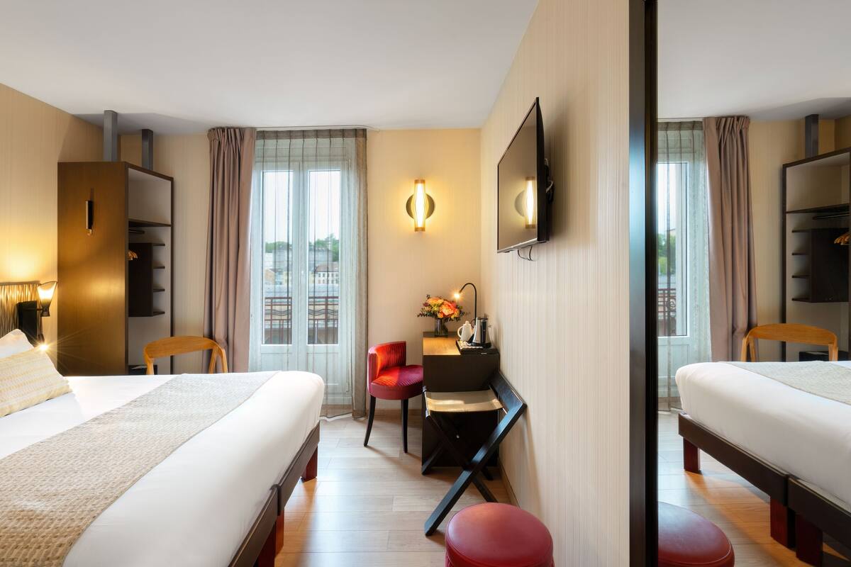 Ein Hotelzimmer mit zwei Betten, einem Schreibtisch, einem Sessel, einem Fernseher und einem großen Fenster mit Vorhängen.
