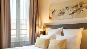 Une chambre d’hôtel avec un grand lit, des rideaux beiges, un tableau abstrait accroché au mur et une vue sur les bâtiments environnants.