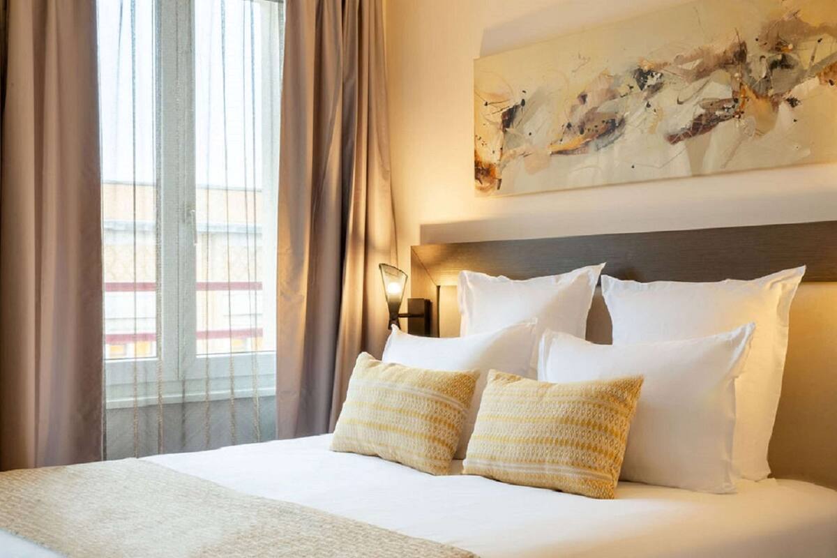 Ein Hotelzimmer mit einem großen Bett, beigen Vorhängen, einem an der Wand hängenden abstrakten Gemälde und Blick auf Gebäude.