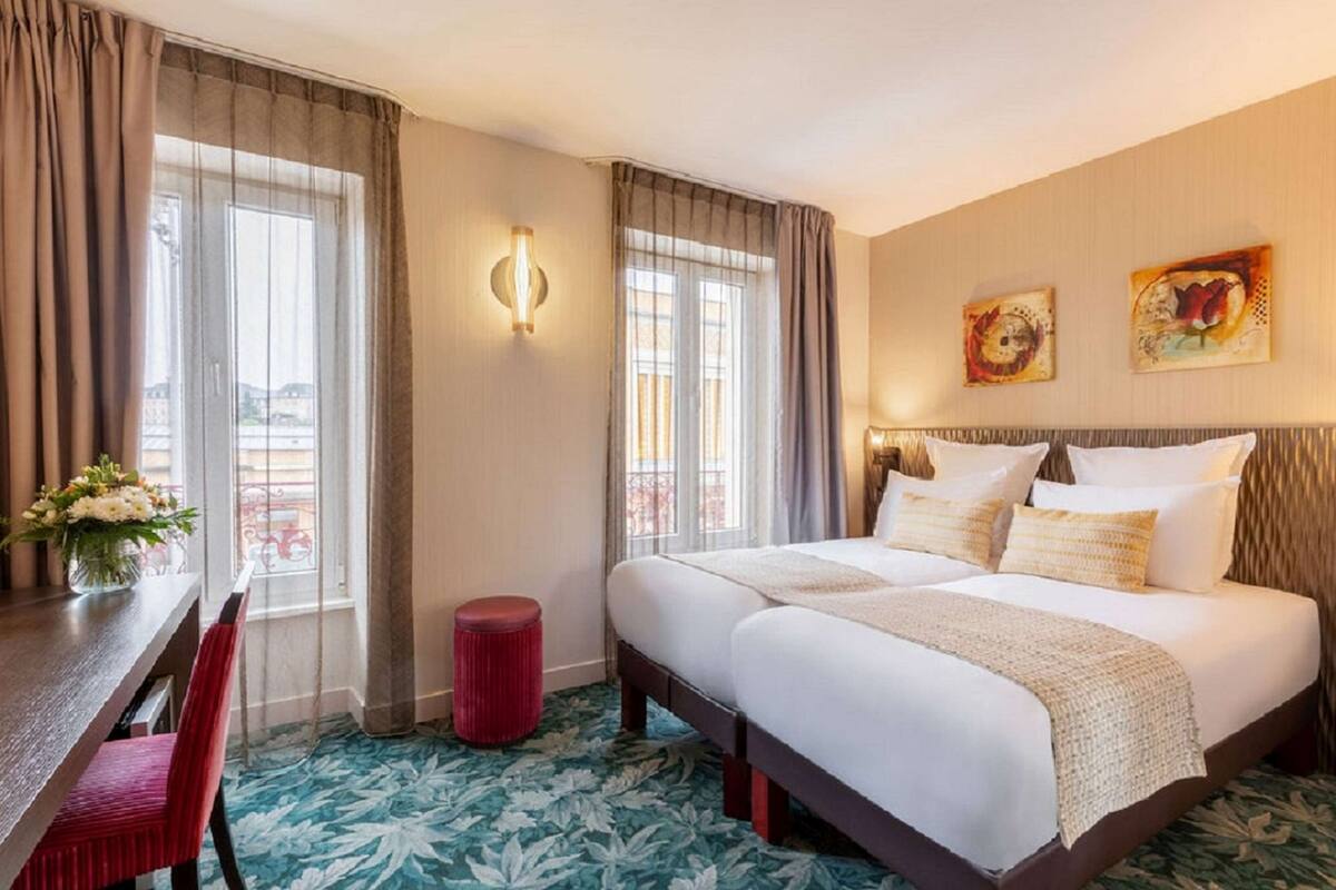 Ein Hotelzimmer mit Doppelbett, zwei Fenstern mit Vorhängen, einem kleinen roten Hocker, einem Tisch mit Blumenvase und Bildern an der Wand.