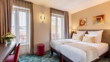 Habitación de hotel con cama doble, dos ventanas con cortinas, un taburete pequeño rojo, una mesa con un jarrón de flores y obras de arte en la pared.