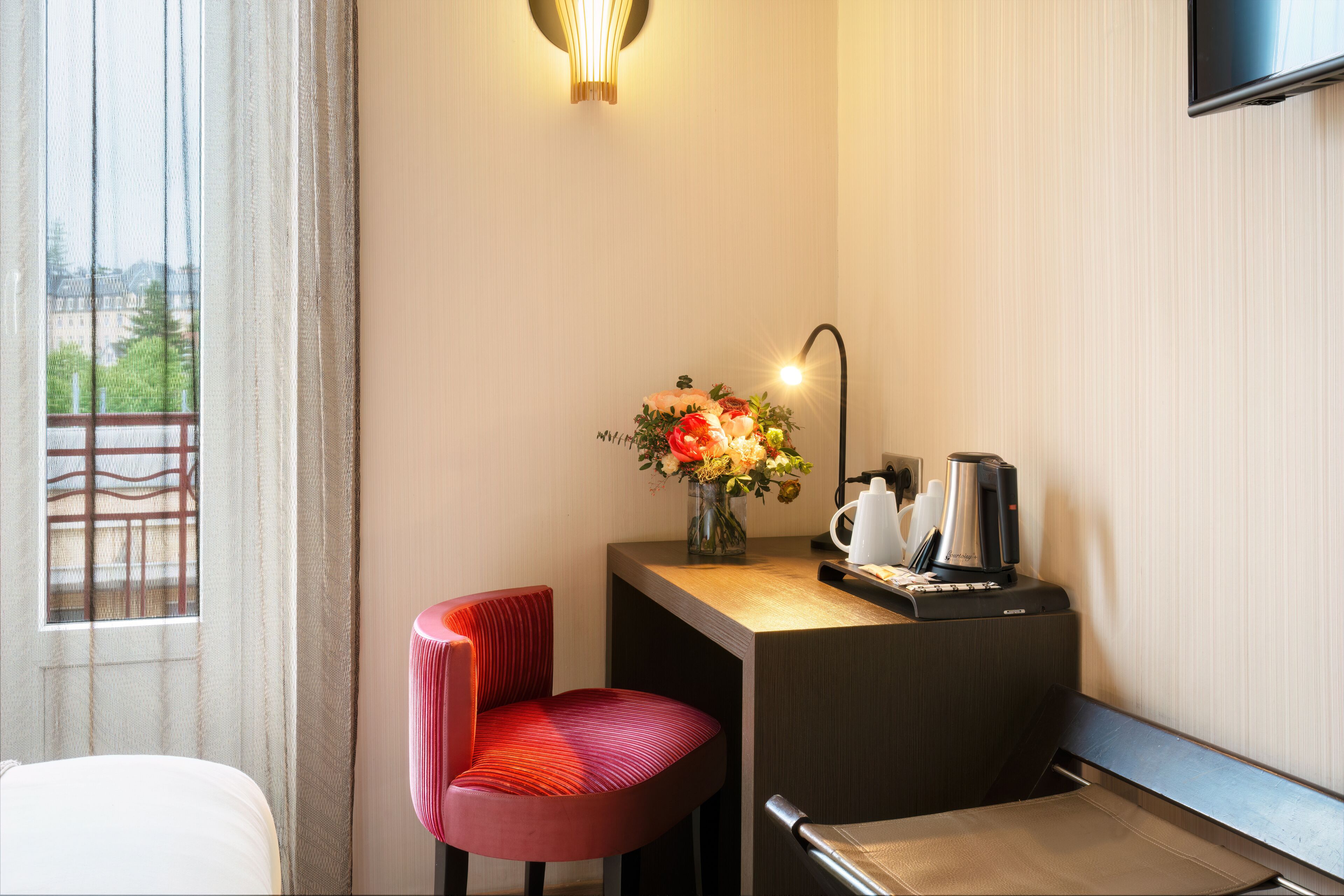 Une chambre d’hôtel avec un lit, un bureau agrémenté d’un vase de fleurs, une machine à café et un téléviseur fixé au mur.