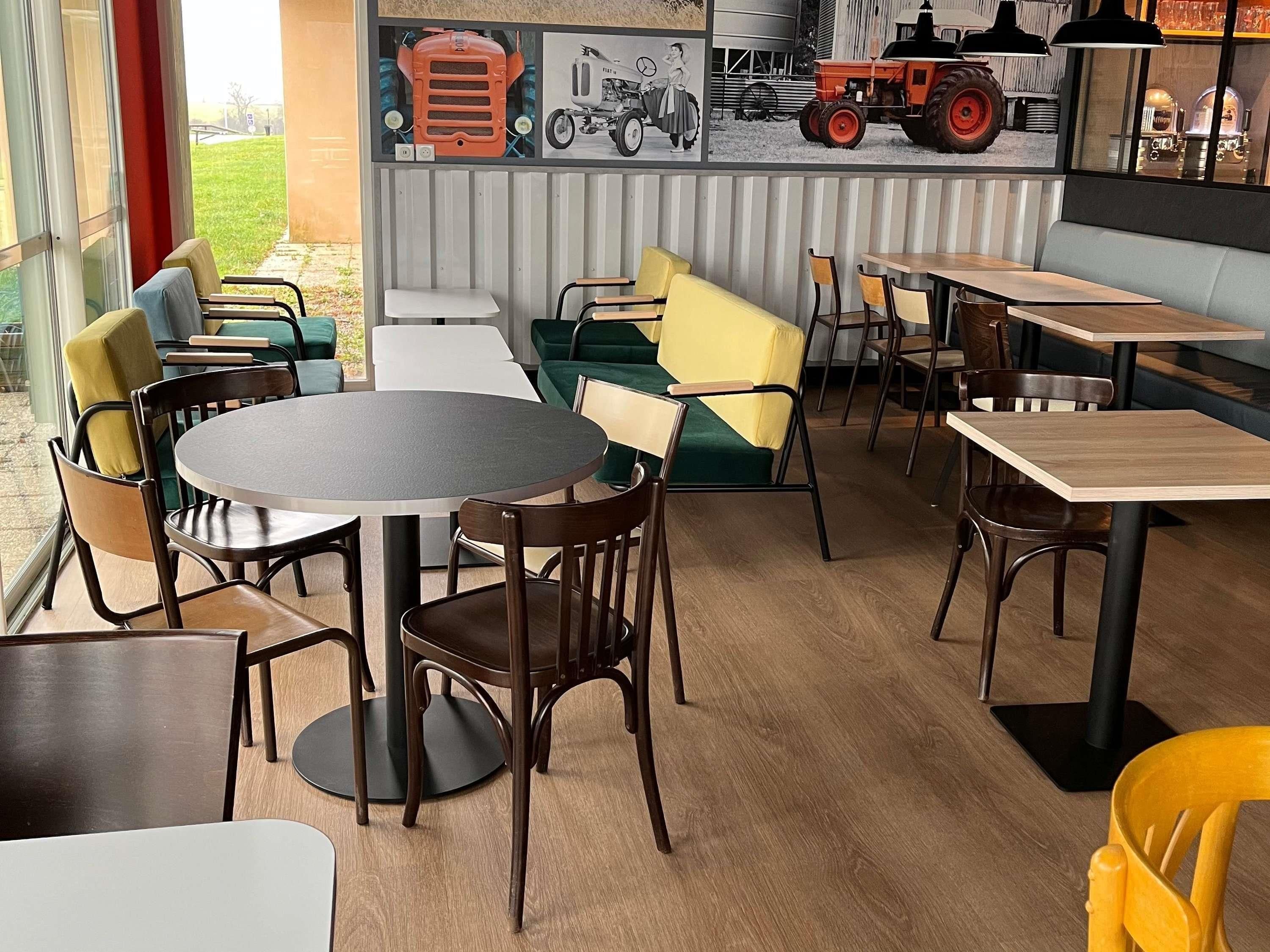 Un restaurant où l’on trouve un mélange de tables rondes et carrées, des sièges rembourrés aux couleurs verte et jaune, et une fresque murale représentant des tracteurs d’époque.