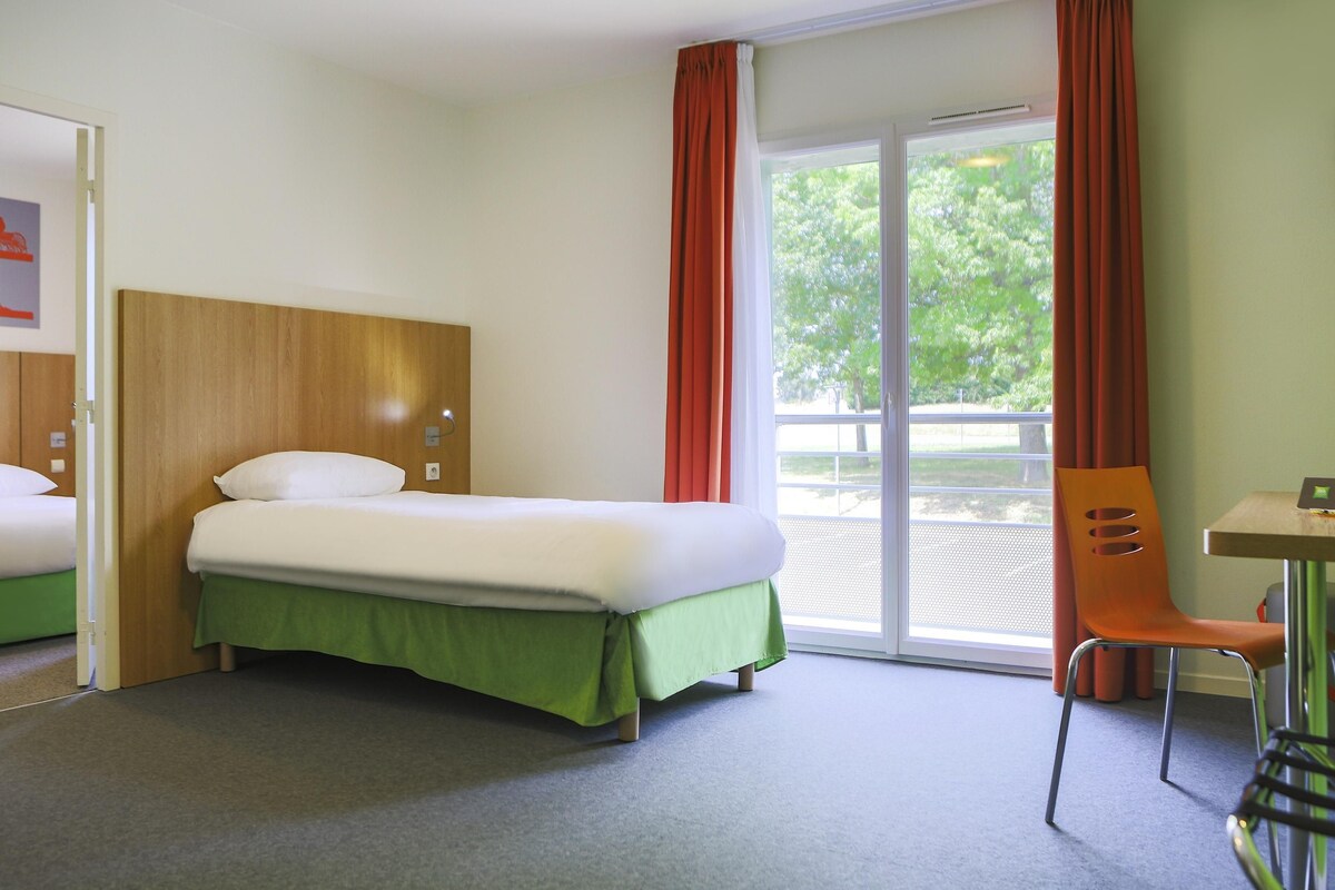 A hotel room with a bed, a chair, a table, and a sliding glass door leading to an outdoor area.