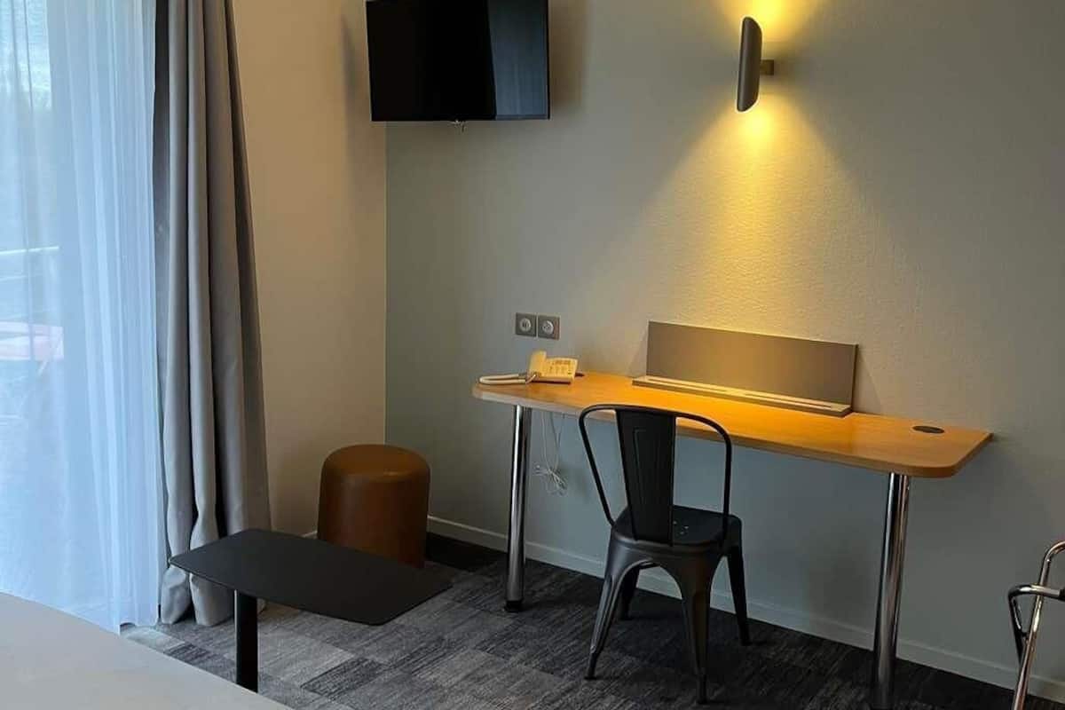A hotel room with a desk, chair, TV, and a small stool.