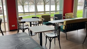 A dining area with tables and chairs, large windows with a view of trees and a body of water.