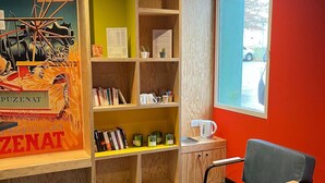 A room with a green chair, wooden shelves with books and a menu, a small table with a kettle, and a window with a view of a car.