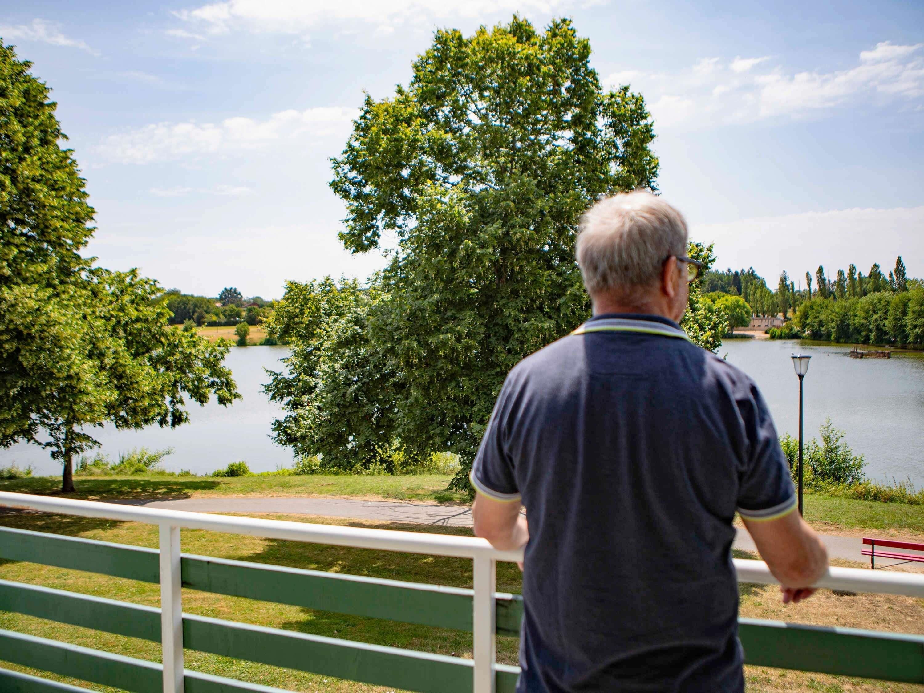 A man standing by a railing overlooking a lake with trees and a distant building.