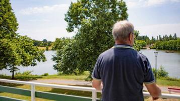 A man standing by a railing overlooking a lake with trees and a distant building.