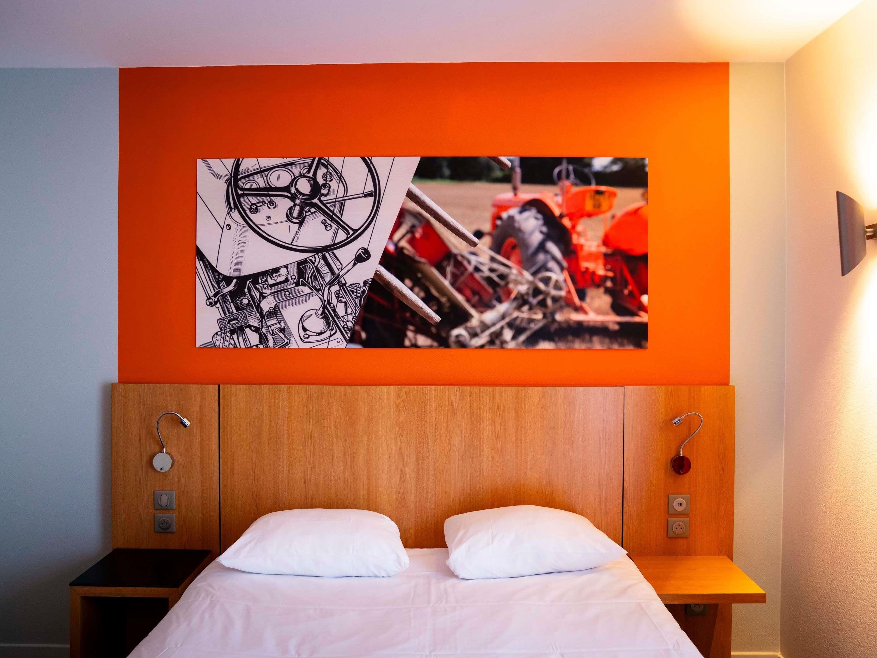 Une chambre d’hôtel comprenant un lit, une tête de lit avec un panneau en bois, une œuvre d’art fixée au mur représentant des machines, et une table de chevet avec une lampe.