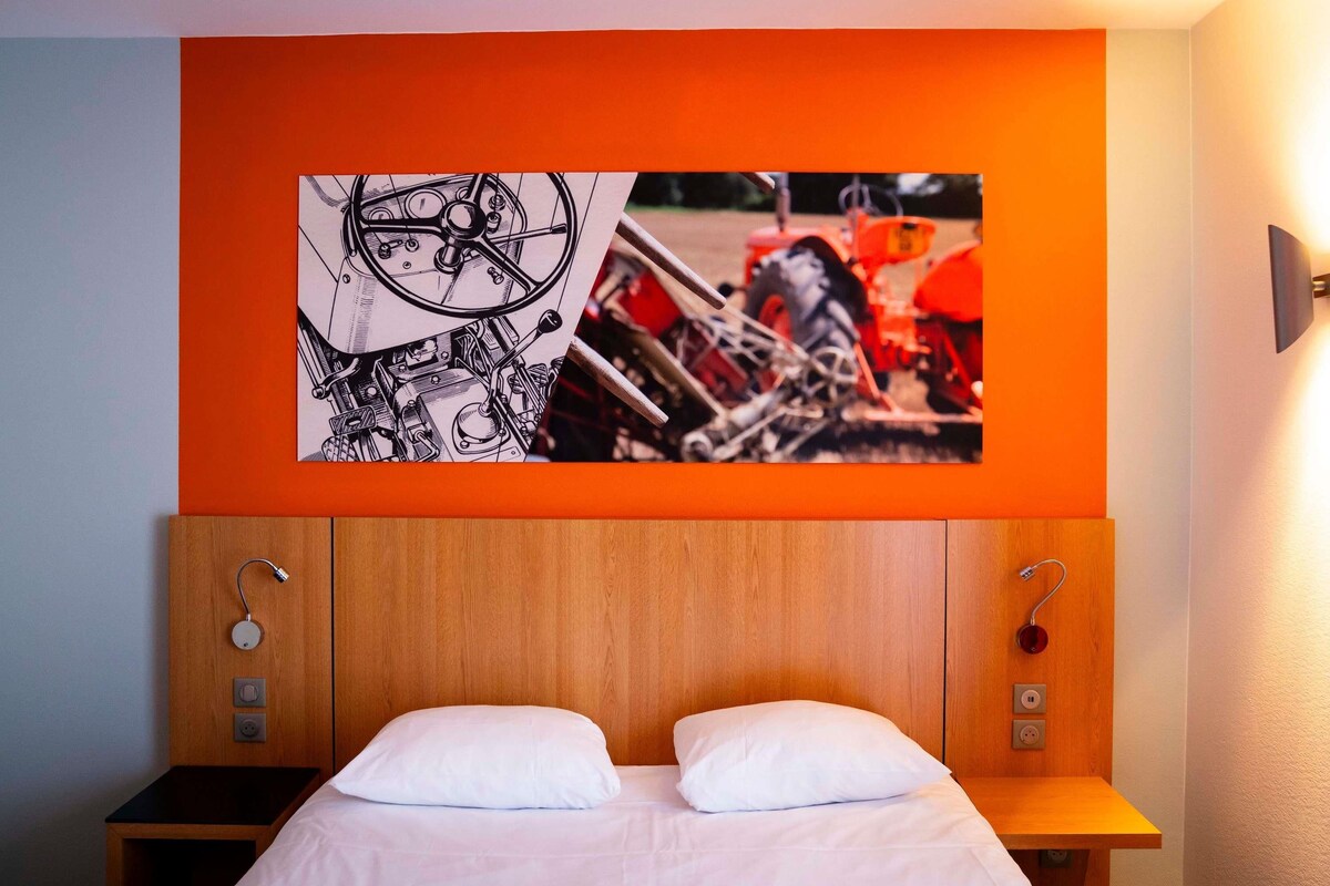 A hotel room with a bed, a headboard with a wooden panel, a wall-mounted artwork depicting machinery, and a bedside table with a lamp.