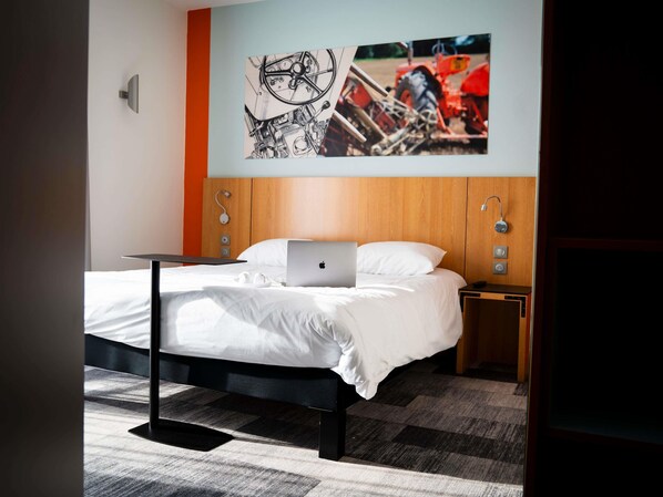 A hotel room with a bed, a laptop, a bedside table, a wall-mounted light, and a wall art piece.