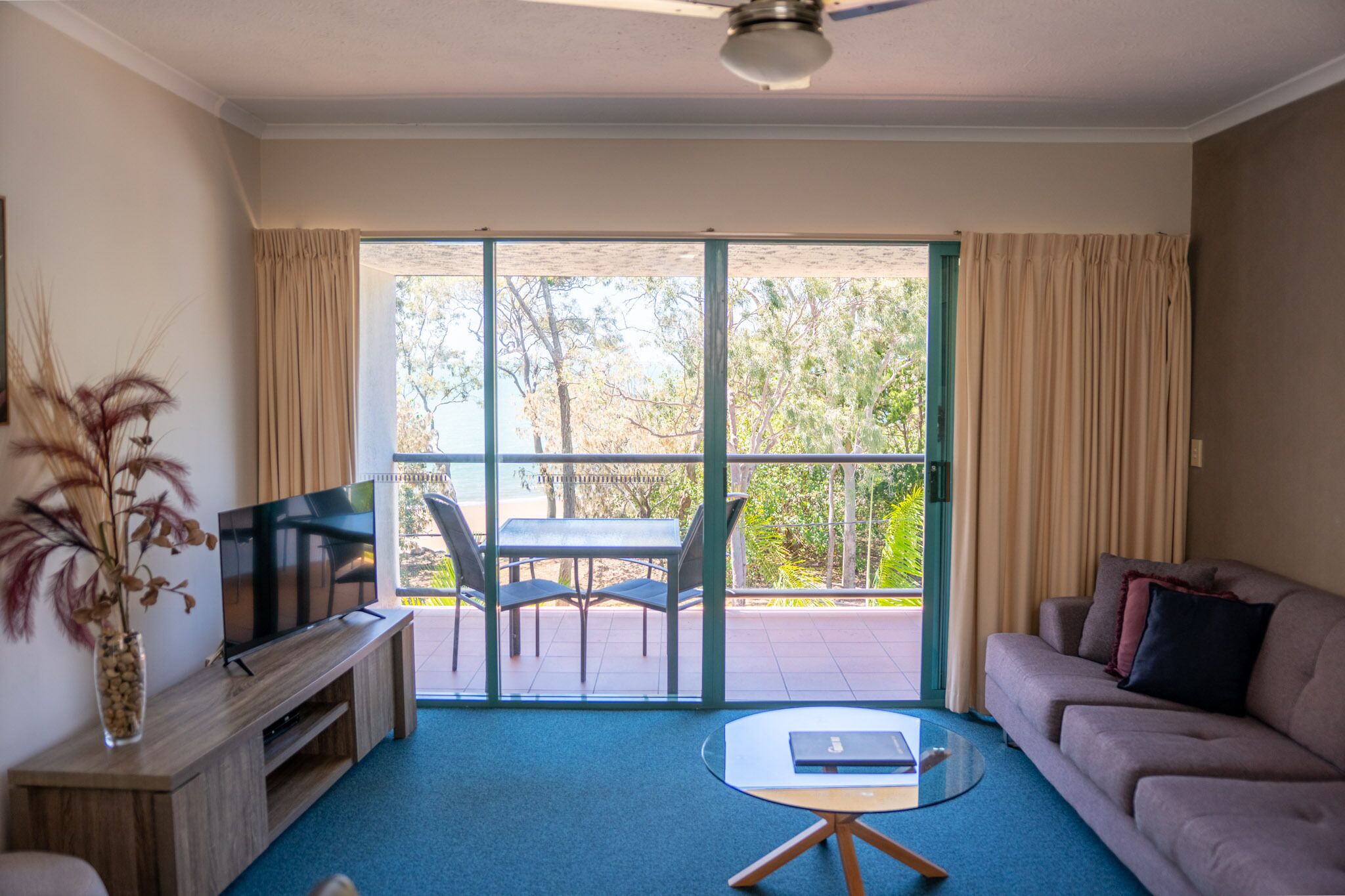 A living room with a TV, sofa, and a table. There is a balcony with a table and chairs, and a view of trees and a building.