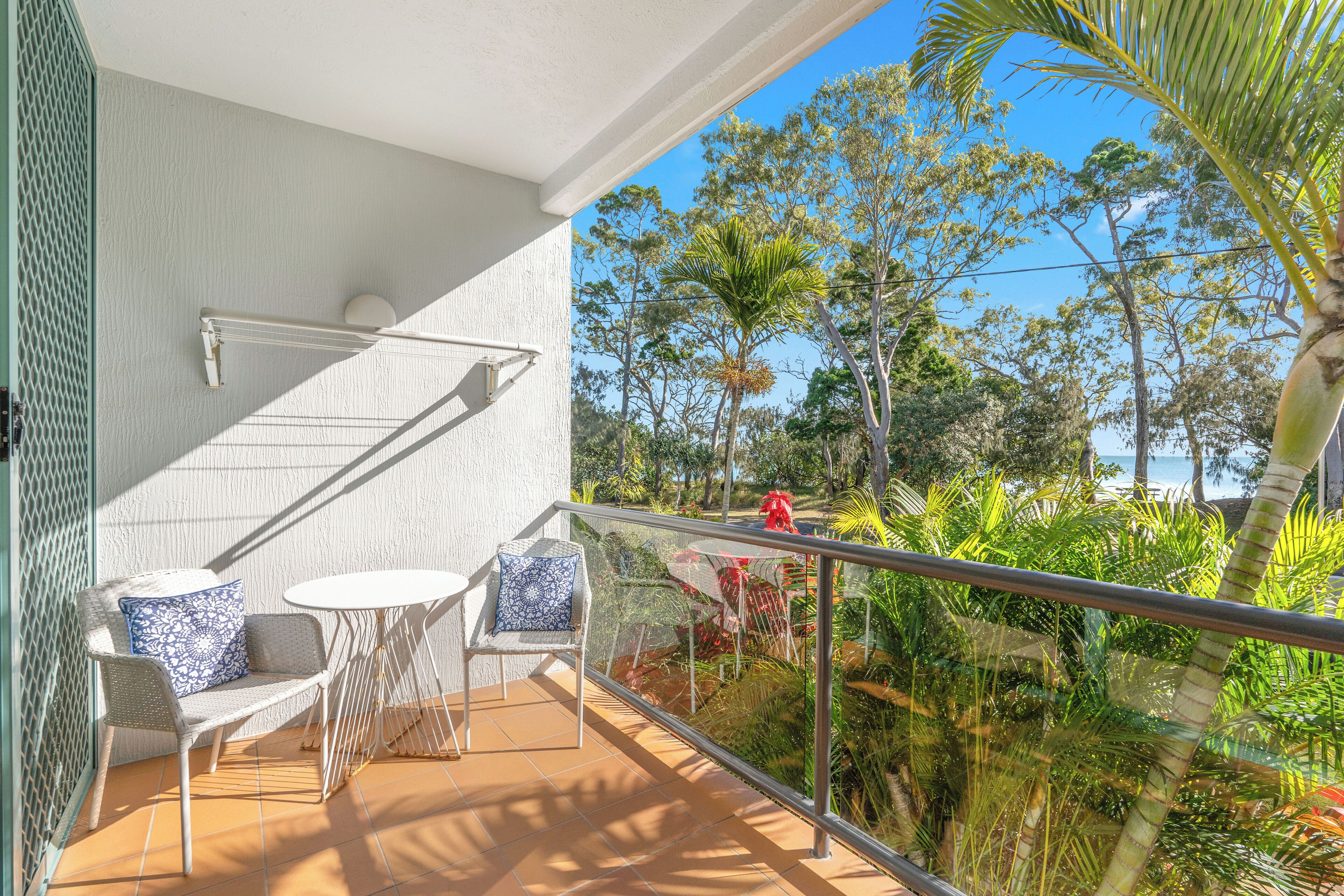 A balcony with a white chair and table, a metal railing, and a view of trees and a beach.