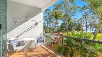 A balcony with a white chair and table, a metal railing, and a view of trees and a beach.