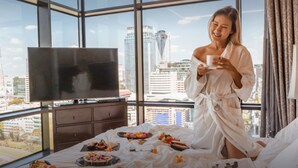 A hotel room with a large window, a flat-screen TV, a bed with a breakfast spread, and a city view. - The St. Regis Bangkok (Bangkok)