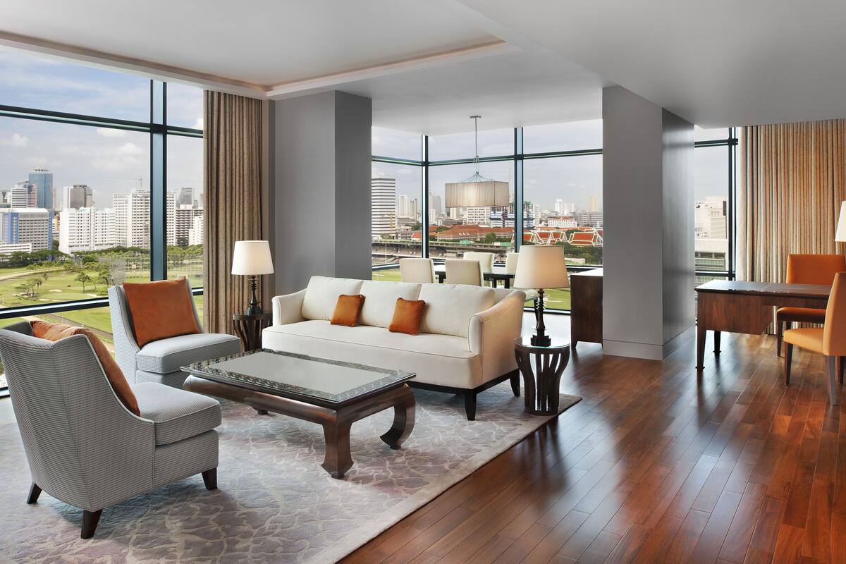 A modern hotel room with a large city view, a sofa, a coffee table, and a dining area.