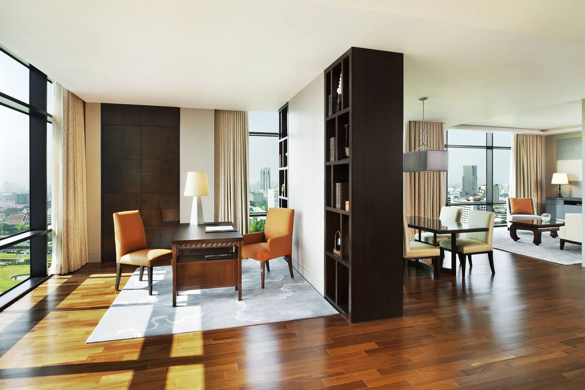A modern hotel room with a large window, a wooden floor, a dining area with a table and chairs, and a bookshelf.