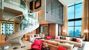 A spacious lobby with a modern staircase, a seating area with red and beige sofas, and large windows offering a city view.