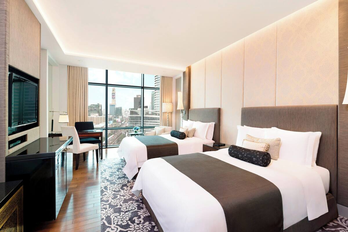 A hotel room with two beds, a desk, a TV, and a large window offering a city view.