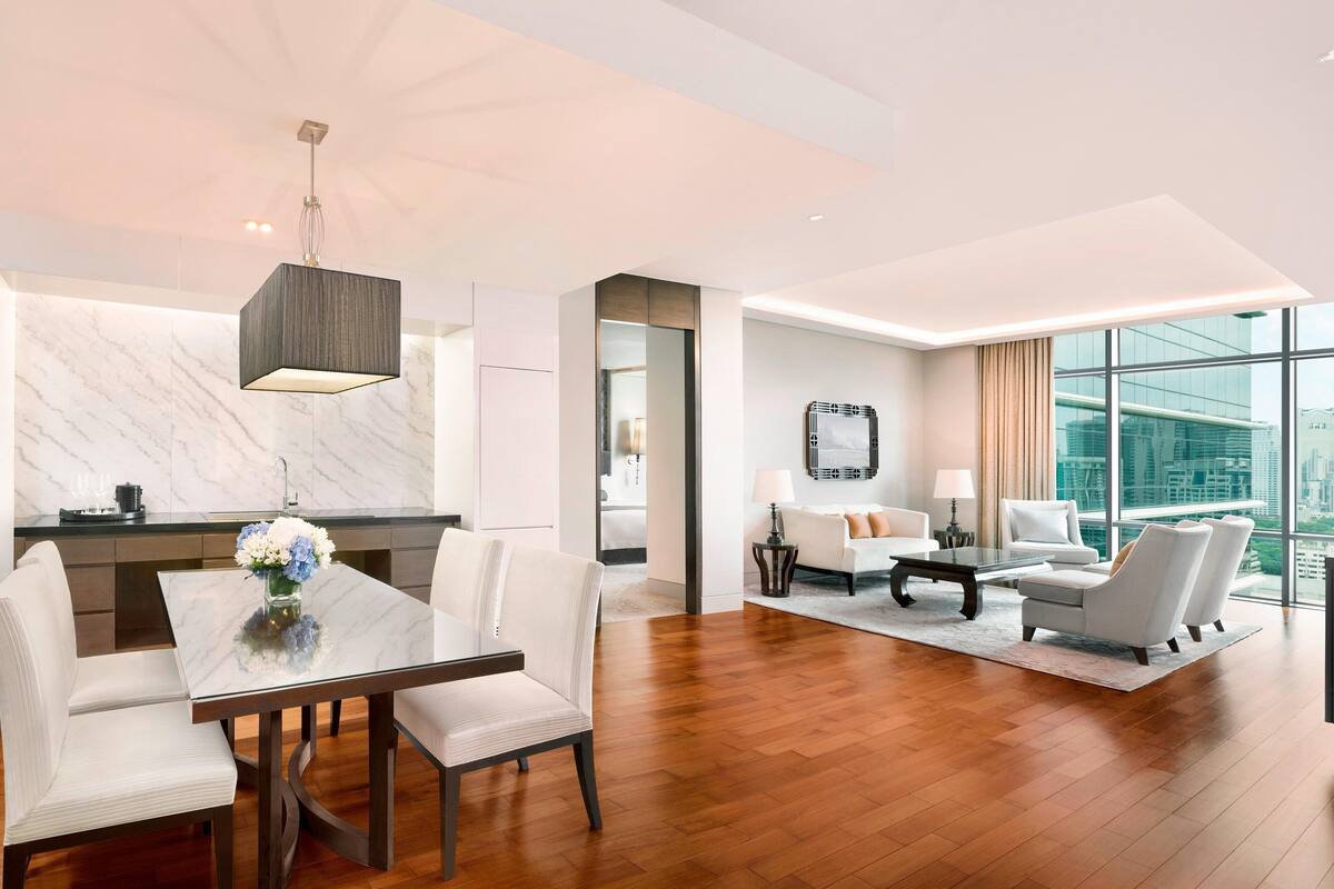 A modern hotel room with a dining area, a living area with a sofa set, and large windows offering a city view.