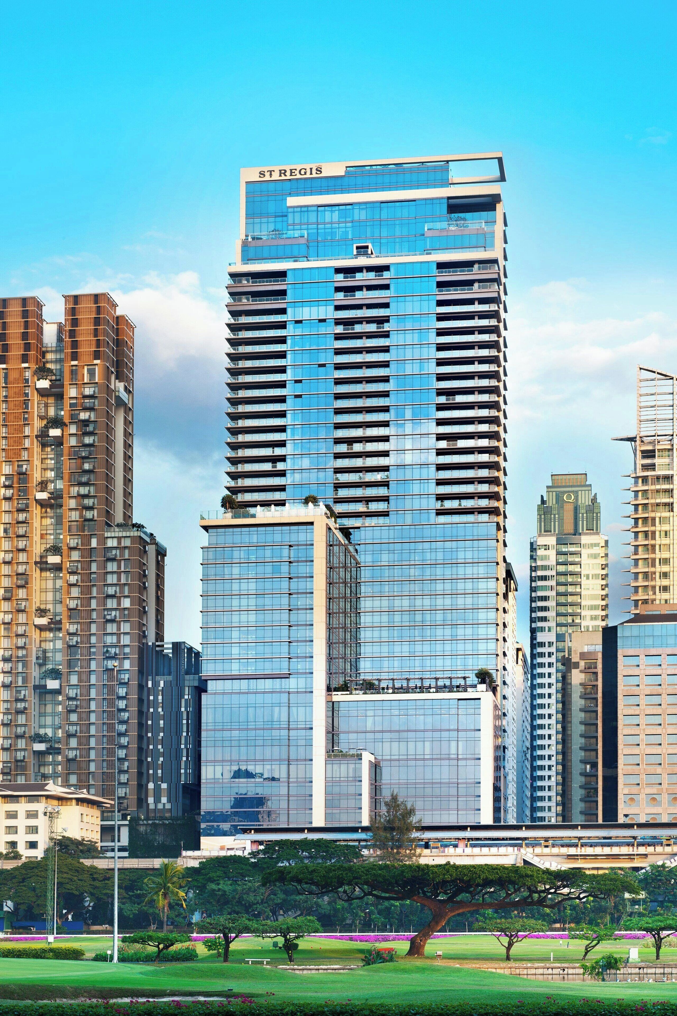 Un moderno edificio de gran altura con fachada de vidrio, rodeado de otros edificios altos y un parque con árboles y césped.