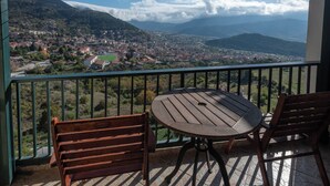 A balcony with a round table and two wooden chairs, overlooking a town and mountains. - Lecadin Hotel (Karpenisi)