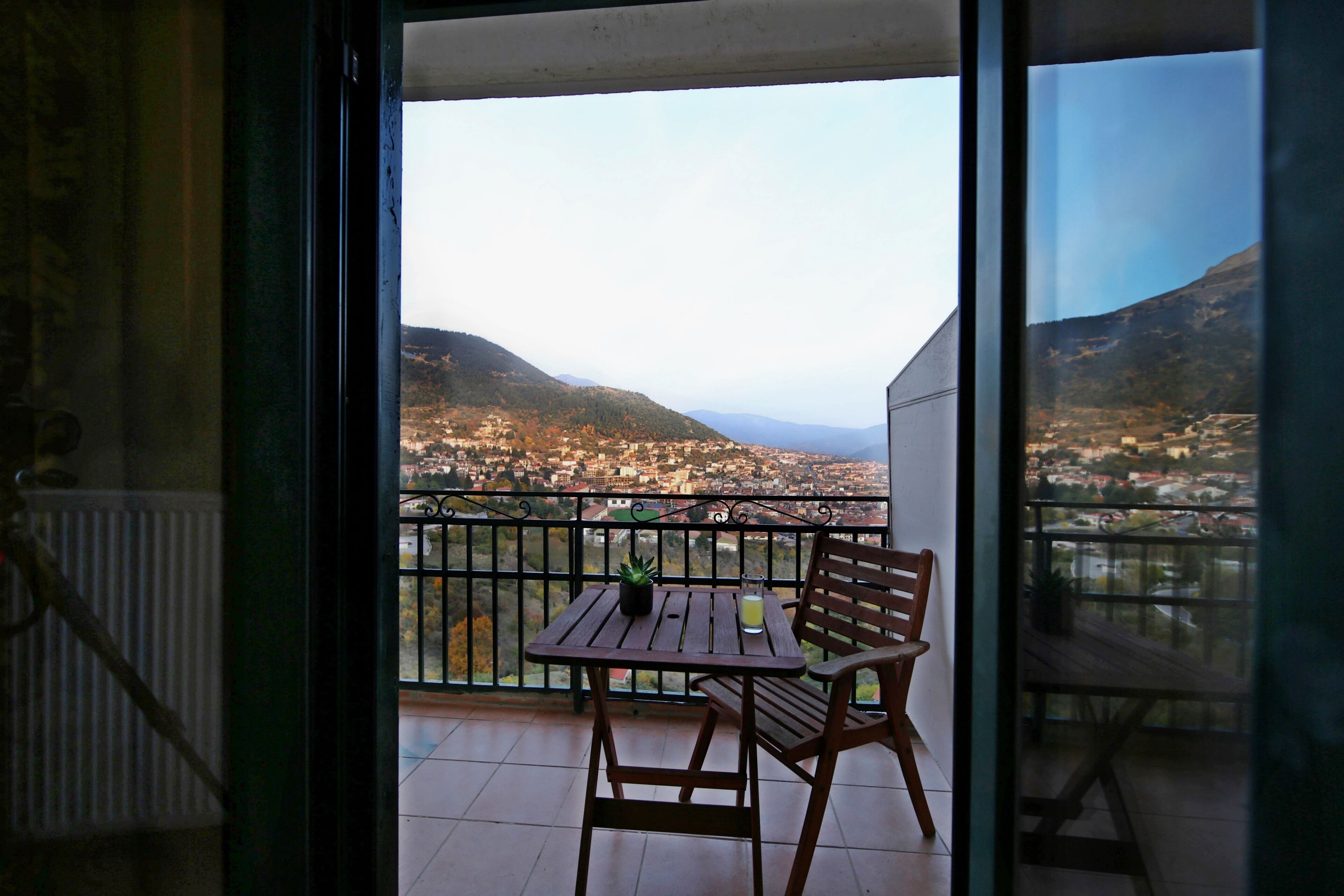 Un balcón con mesa y silla de madera, una planta en maceta y un ventanal con vistas a la ciudad.