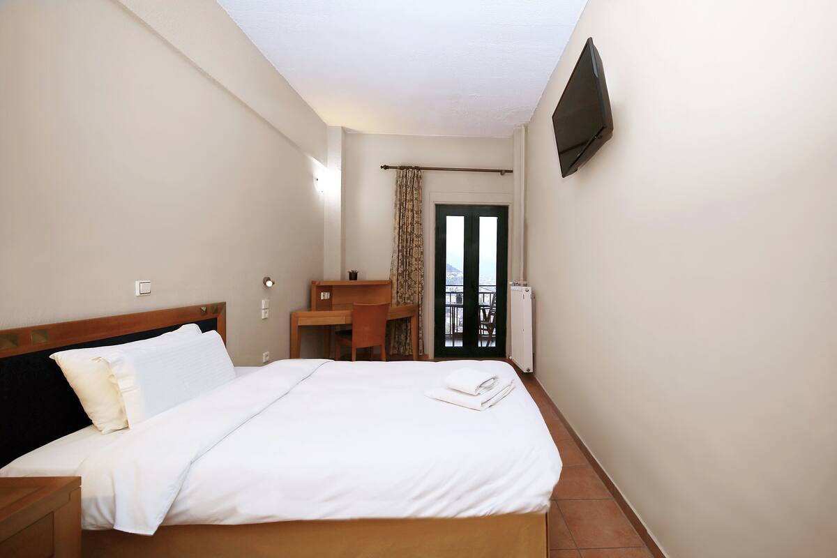 A hotel room with a bed, a desk, a chair, a TV mounted on the wall, and a view of the outside through a door.
