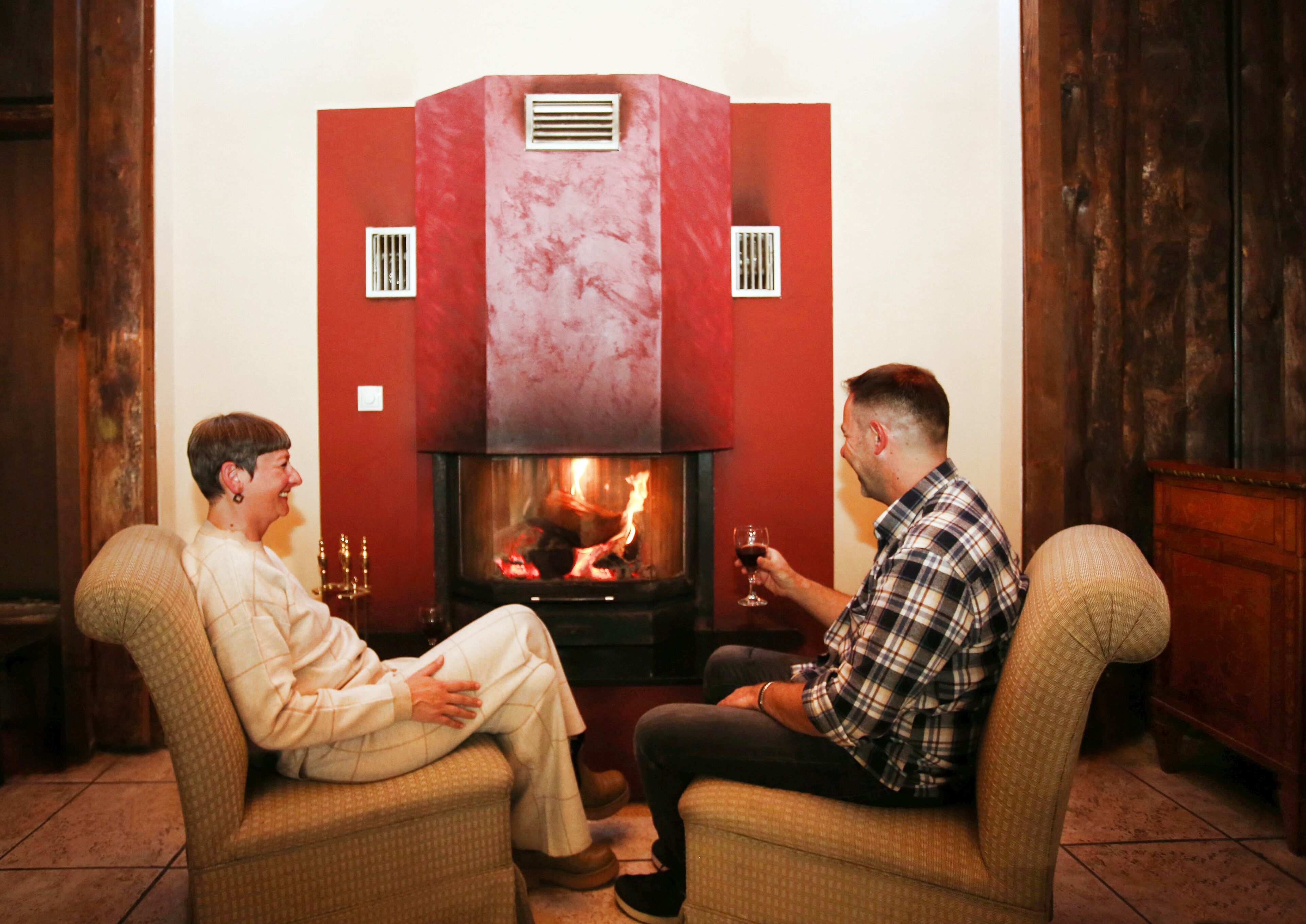 Dos personas sentadas en sillones frente a una chimenea con un marco de mármol rojo y fuego visible.