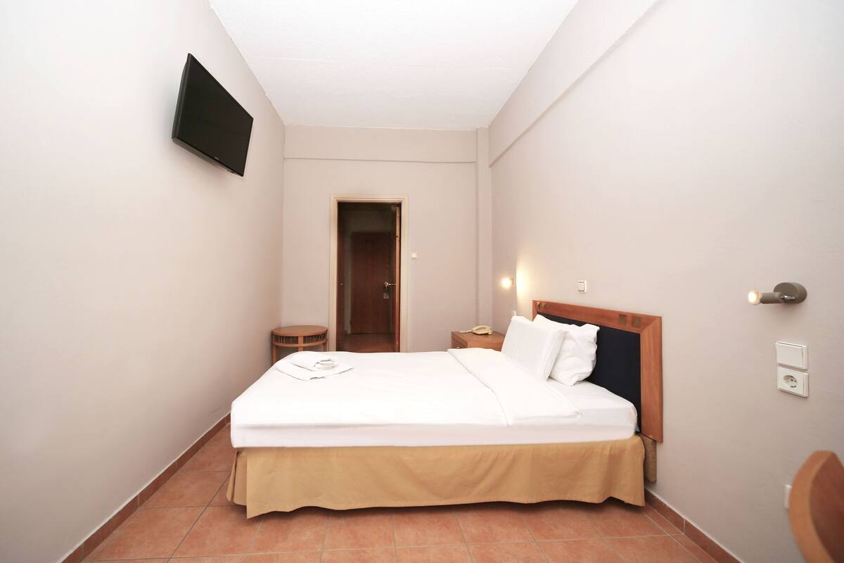 A hotel room with a bed, a bedside table, a TV mounted on the wall, and a door leading to another room.