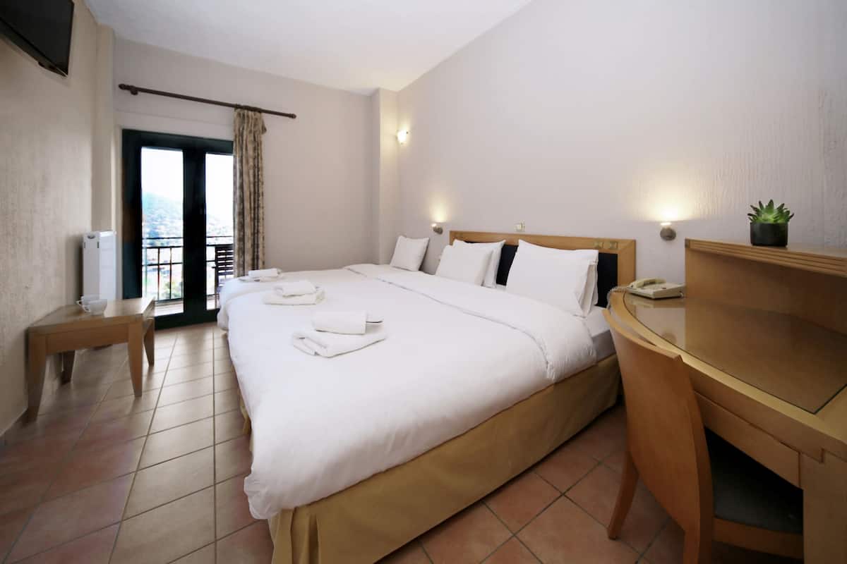 A hotel room with a large bed, a desk, a chair, a small table, a potted plant, and a balcony with a view.