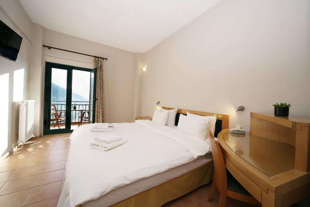 A hotel room with a large bed, a wooden nightstand, a desk, and a view of the outdoors through a sliding door.