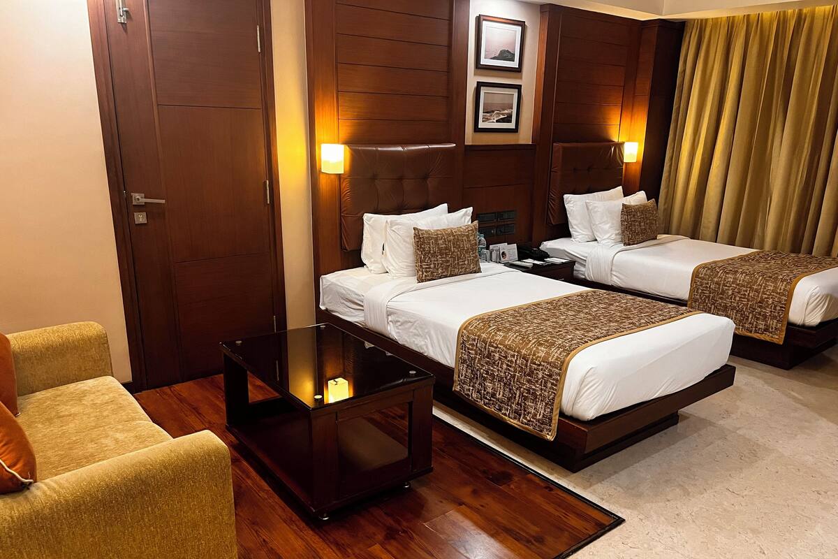 A hotel room with two beds, a small table, a chair, wooden flooring, and wall-mounted lamps.