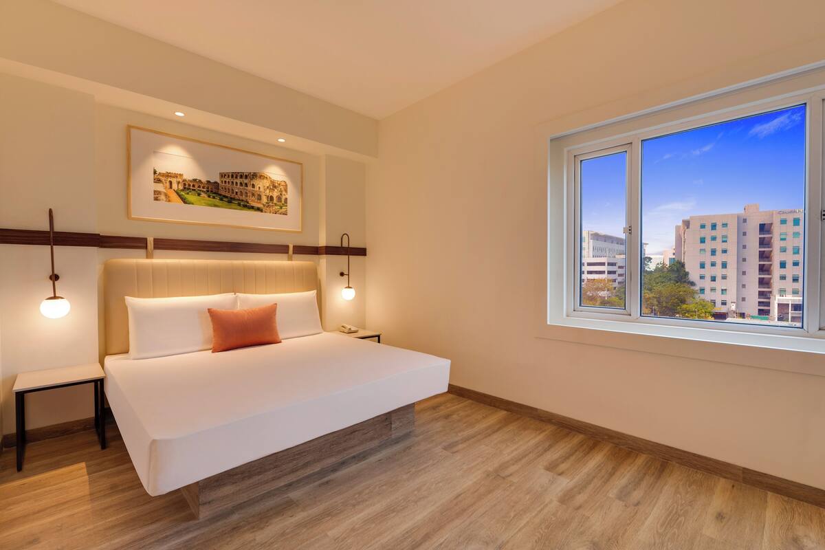 Una habitación de hotel moderna con una cama grande, una mesita de noche, un cuadro enmarcado en la pared y una ventana con vistas a la ciudad.