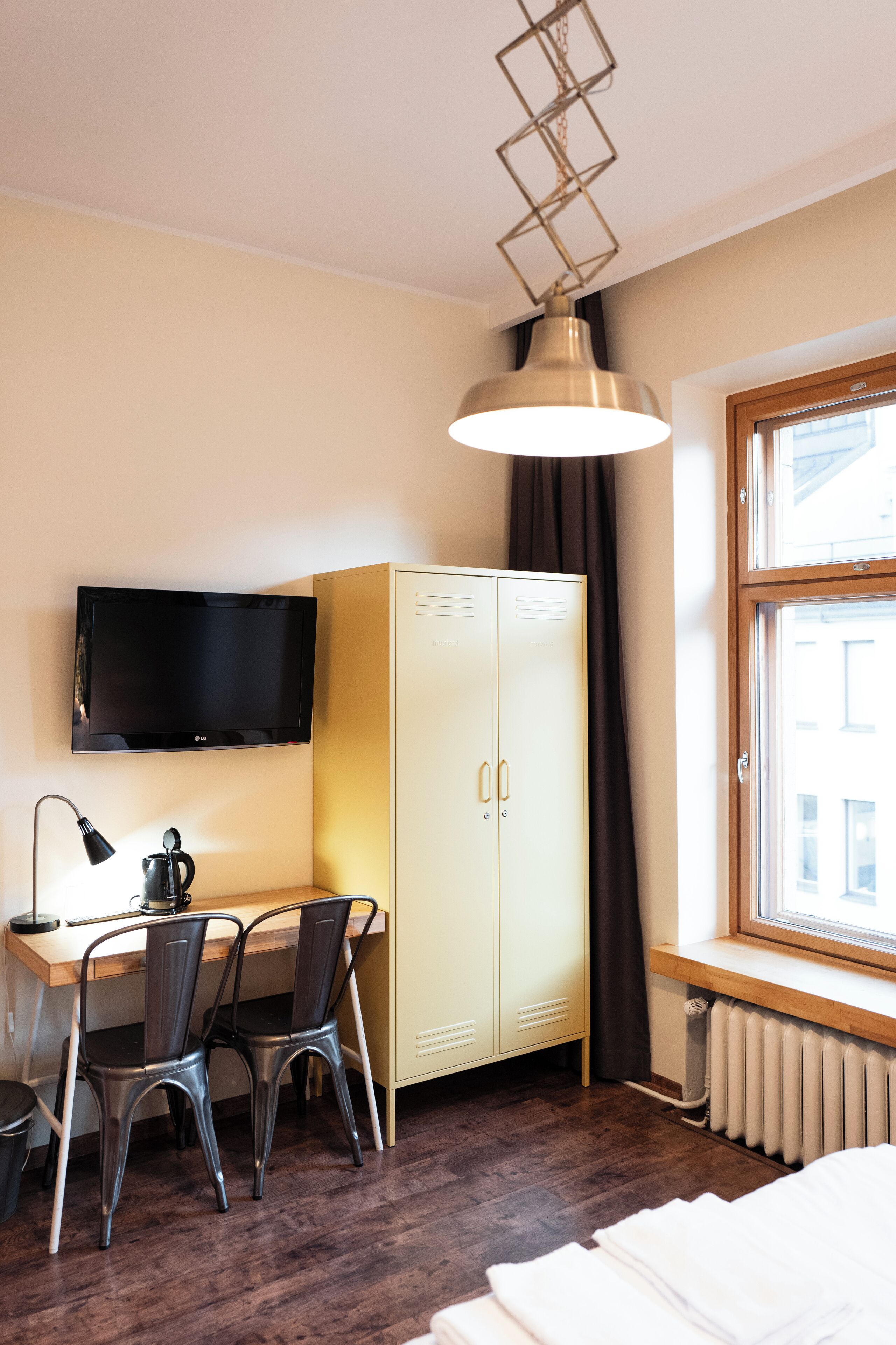 Une pièce comprenant un bureau en bois, deux chaises, un téléviseur, une armoire et une fenêtre donnant sur des immeubles.